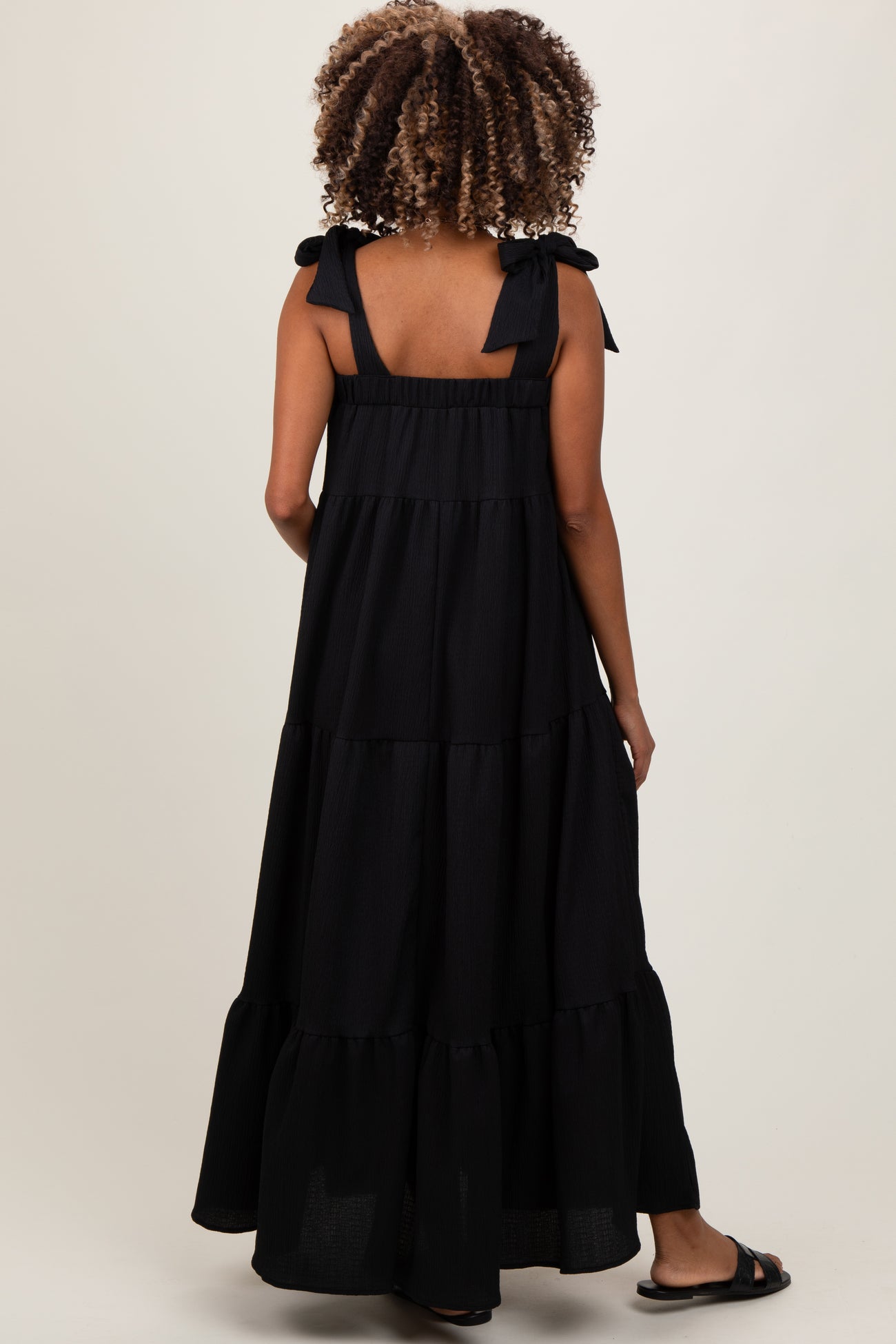 woman wearing a long black maternity dress with tiered design and bow straps, showing her pregnant belly from the back