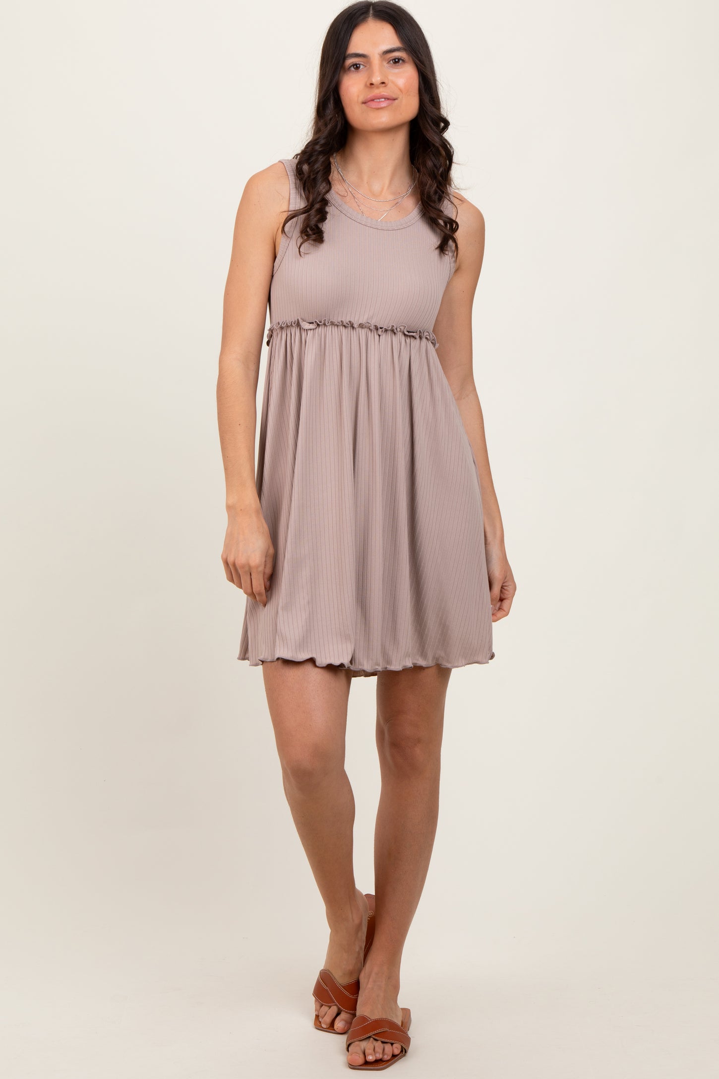 woman wearing a taupe sleeveless dress with a ruched waistline and a flowy skirt, standing and smiling