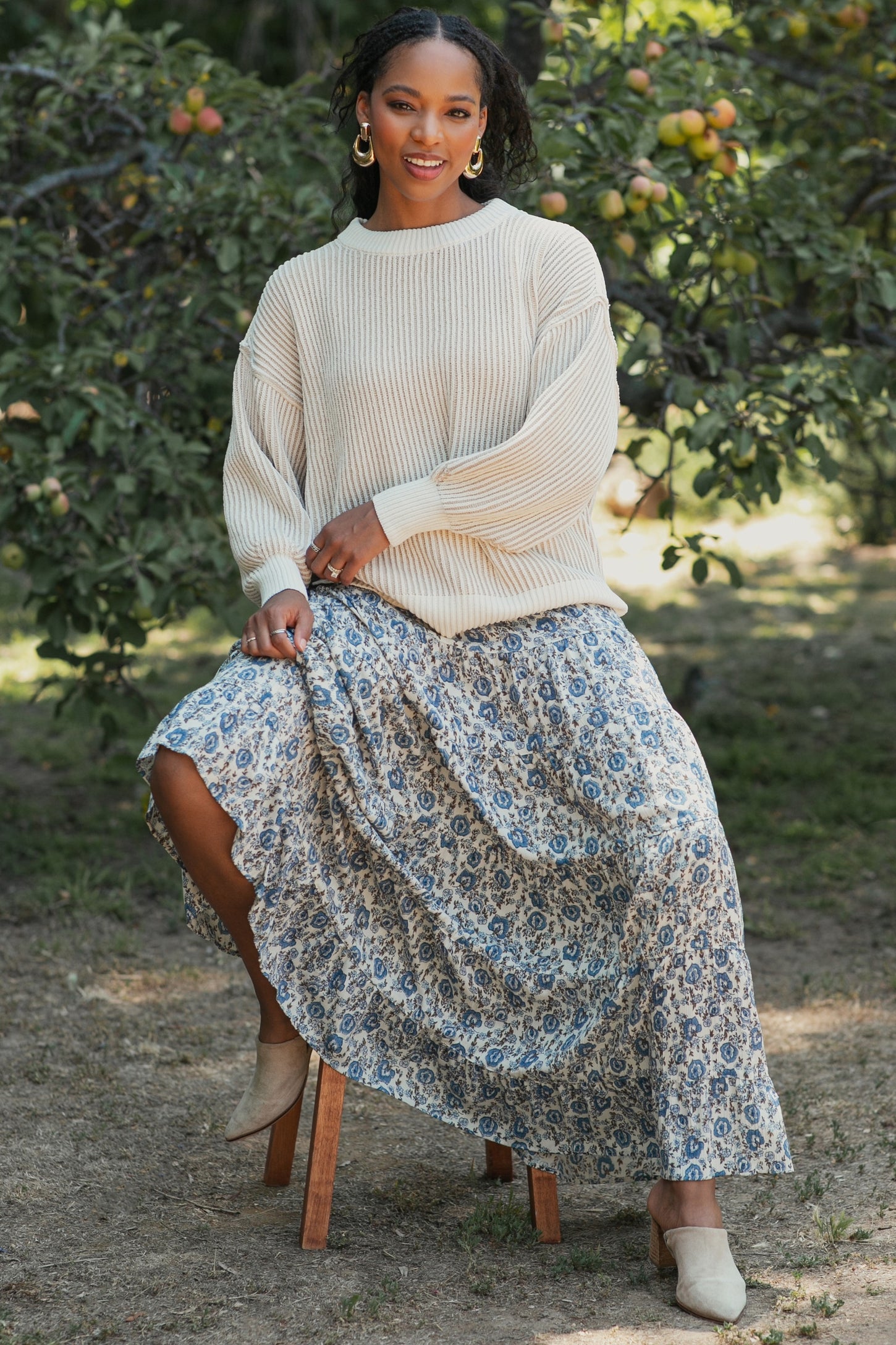 woman wearing a cream oversized sweater and a flowy floral maxi skirt, sitting on a wooden stool outdoors
