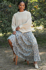 woman wearing a cream oversized sweater and a flowy floral maxi skirt, sitting on a wooden stool outdoors