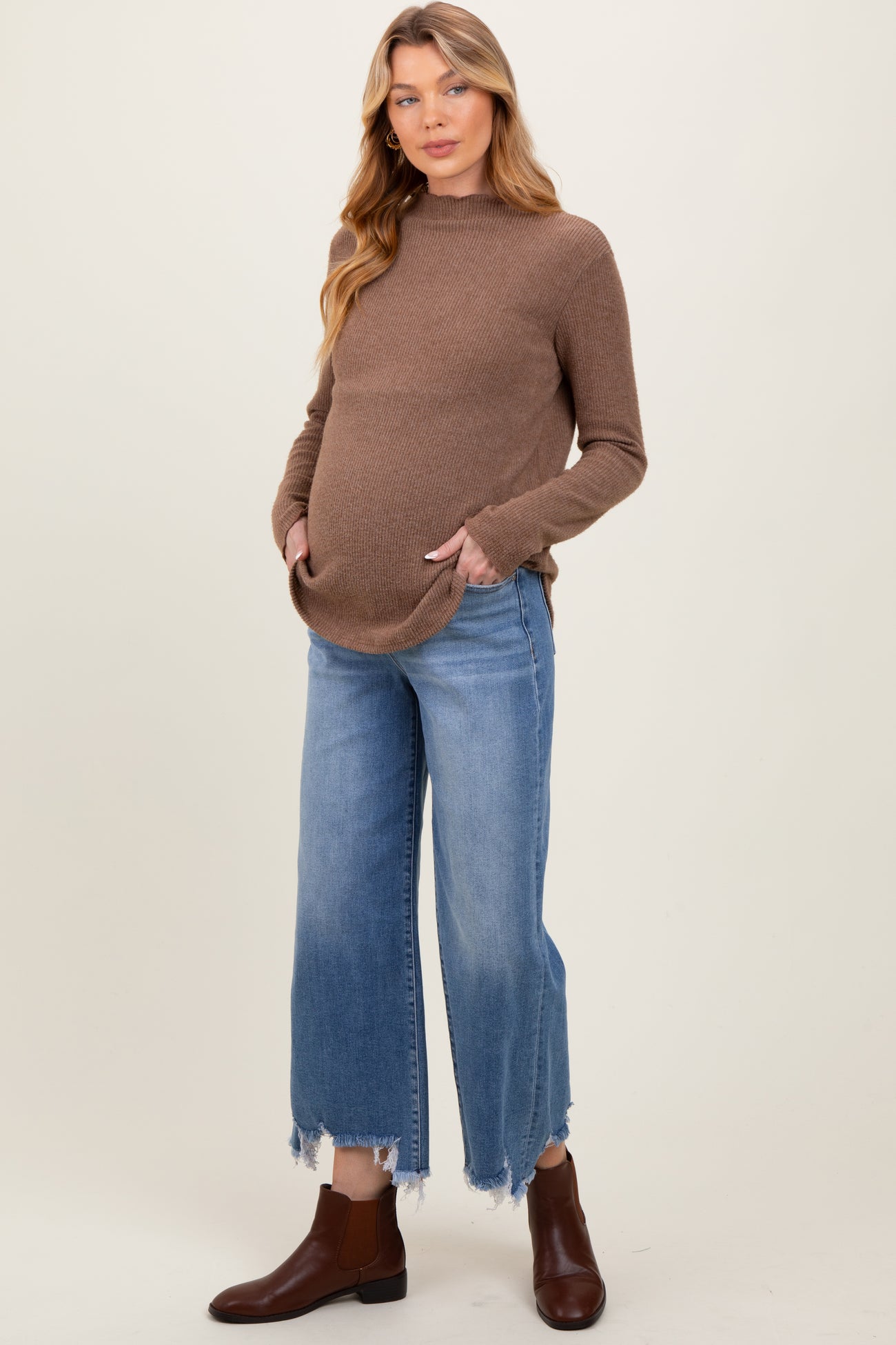 woman wearing a brown maternity sweater with pockets and blue frayed hem denim jeans, showing her baby bump