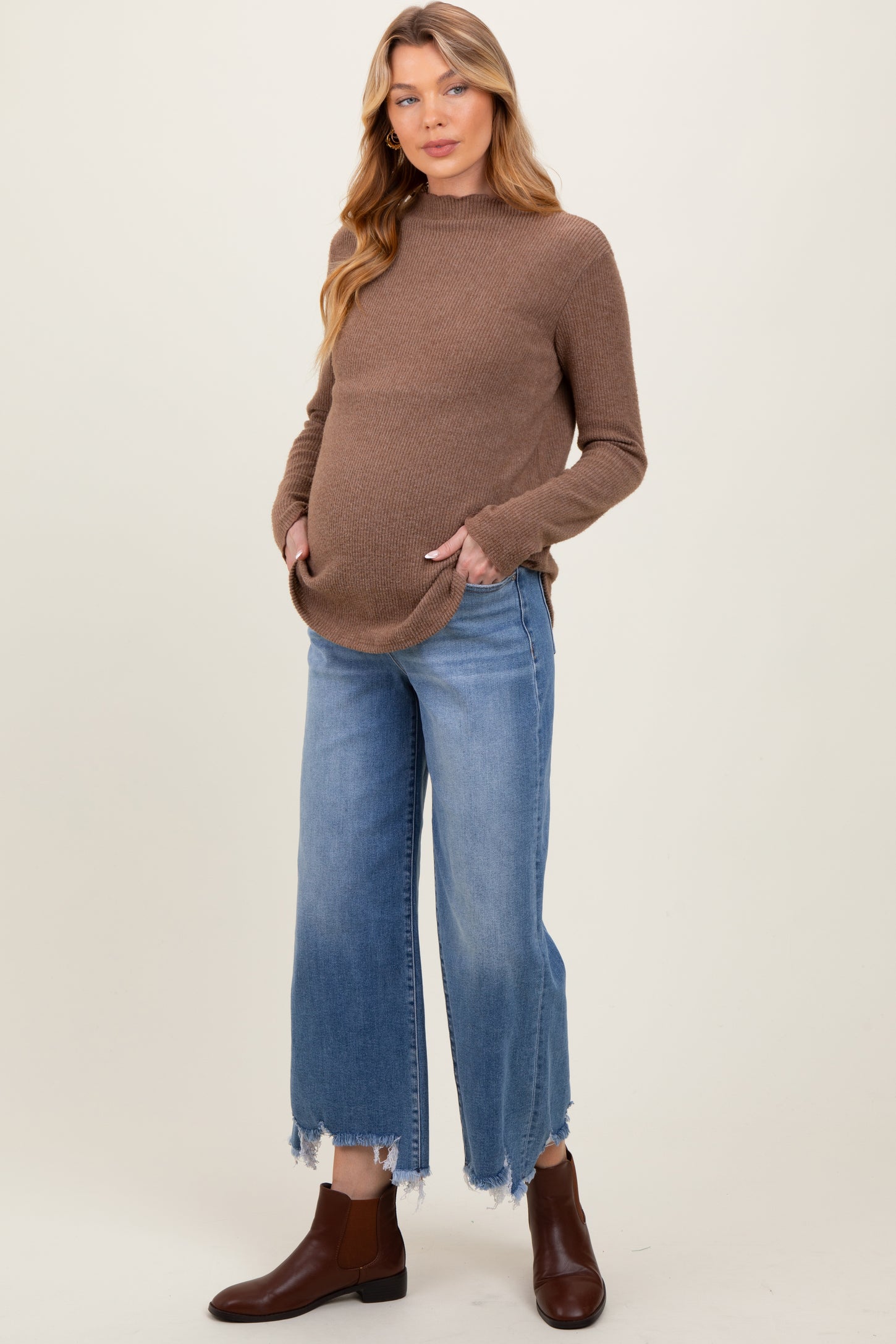 woman wearing a brown maternity sweater with pockets and blue frayed hem denim jeans, showing her baby bump