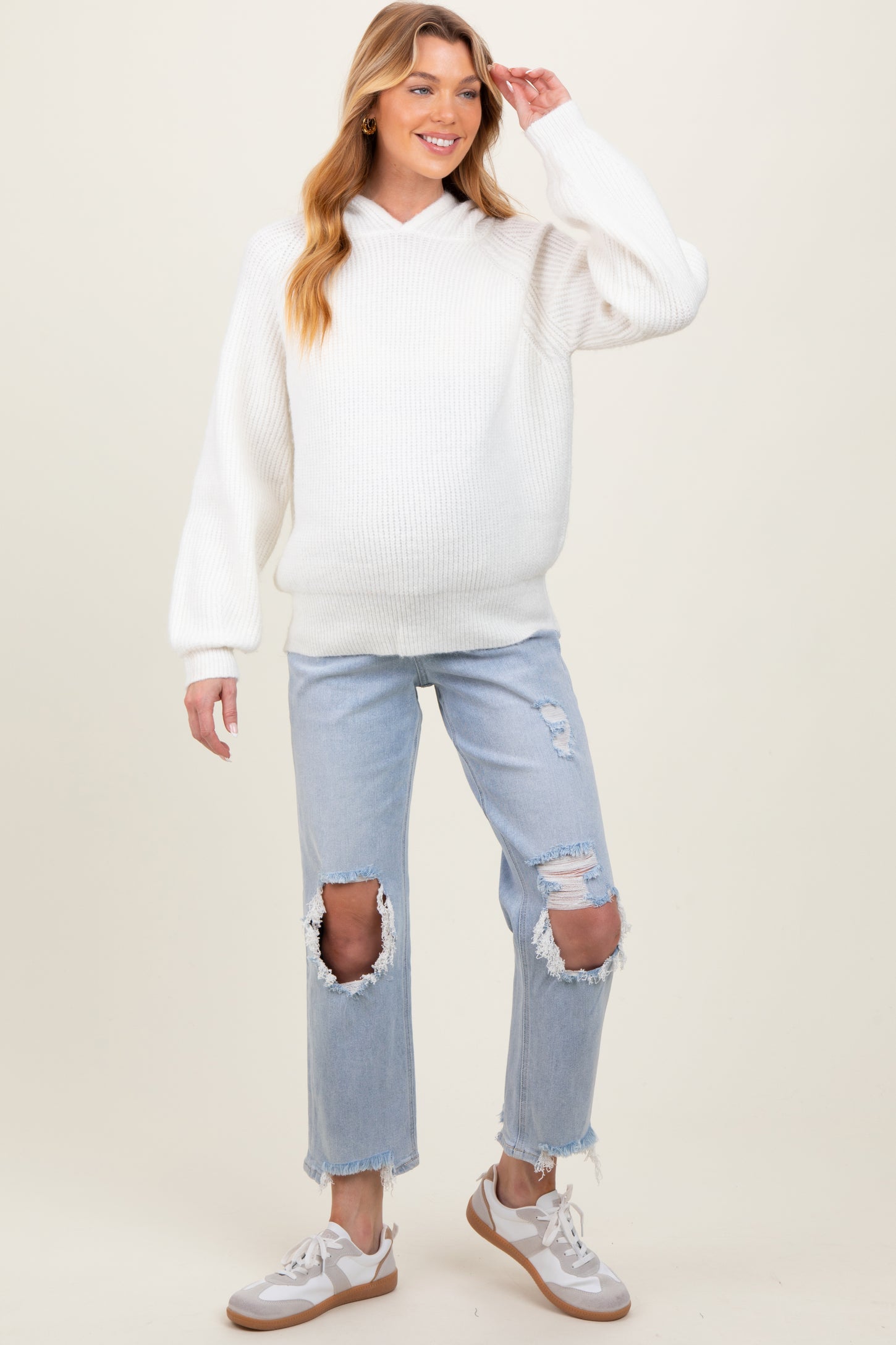 woman wearing a white knit maternity hoodie and distressed light blue jeans, gently touching her baby bump
