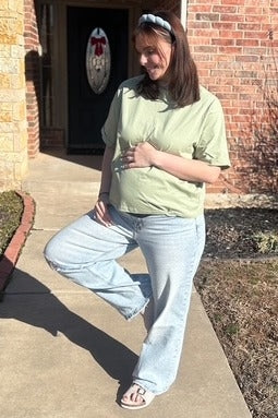 woman wearing a light green maternity t-shirt and light blue wide-leg jeans, holding her baby bump.