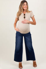 woman wearing a beige short sleeve maternity top and blue wide-leg jeans, holding her baby bump