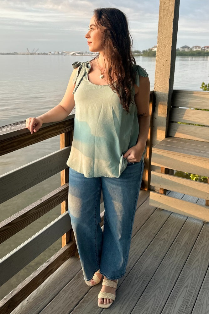 woman wearing a light green maternity tank top with tie straps and blue wide-leg jeans, standing by the water