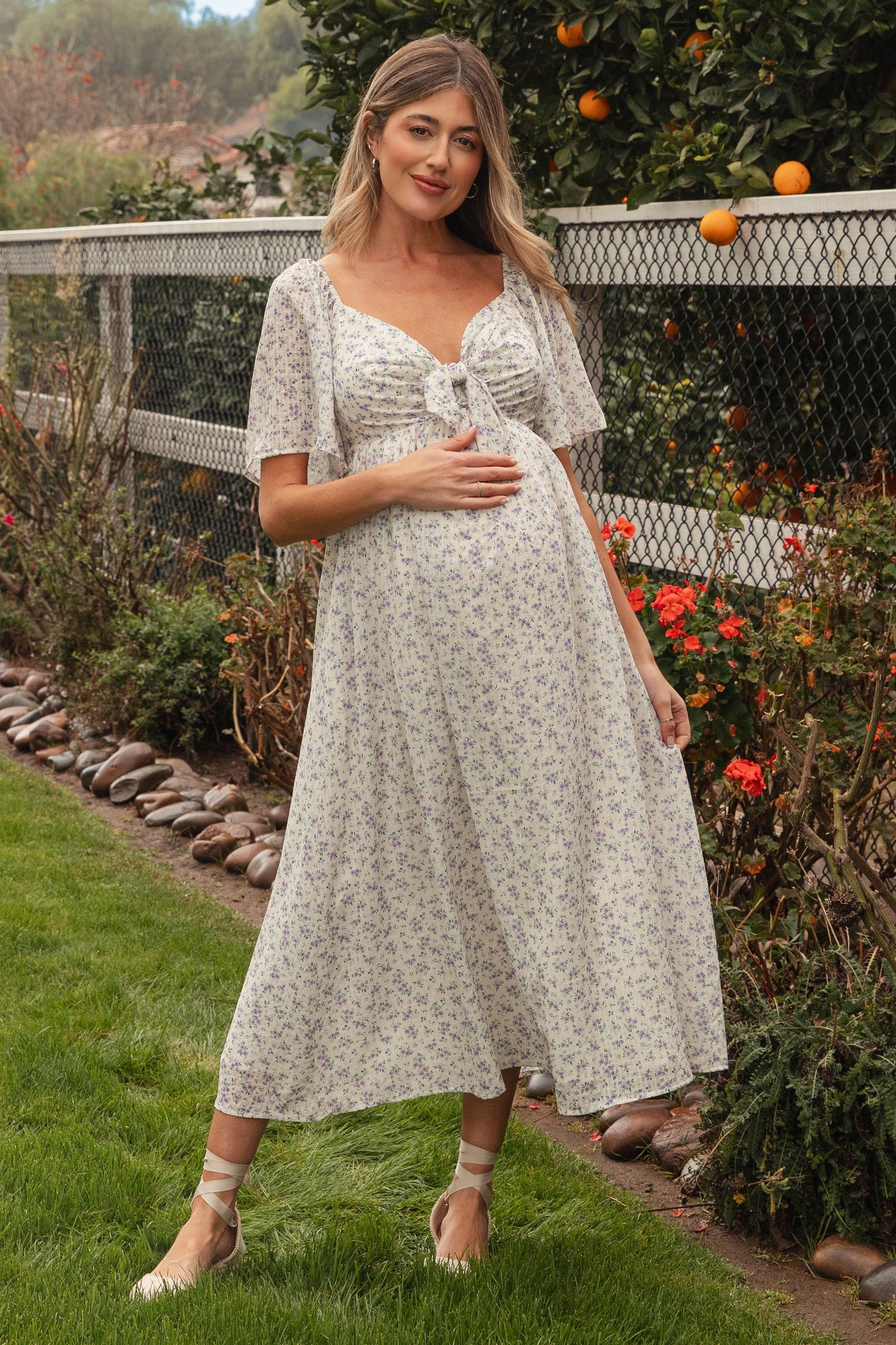woman wearing a white floral maternity dress with short flutter sleeves and holding her baby bump