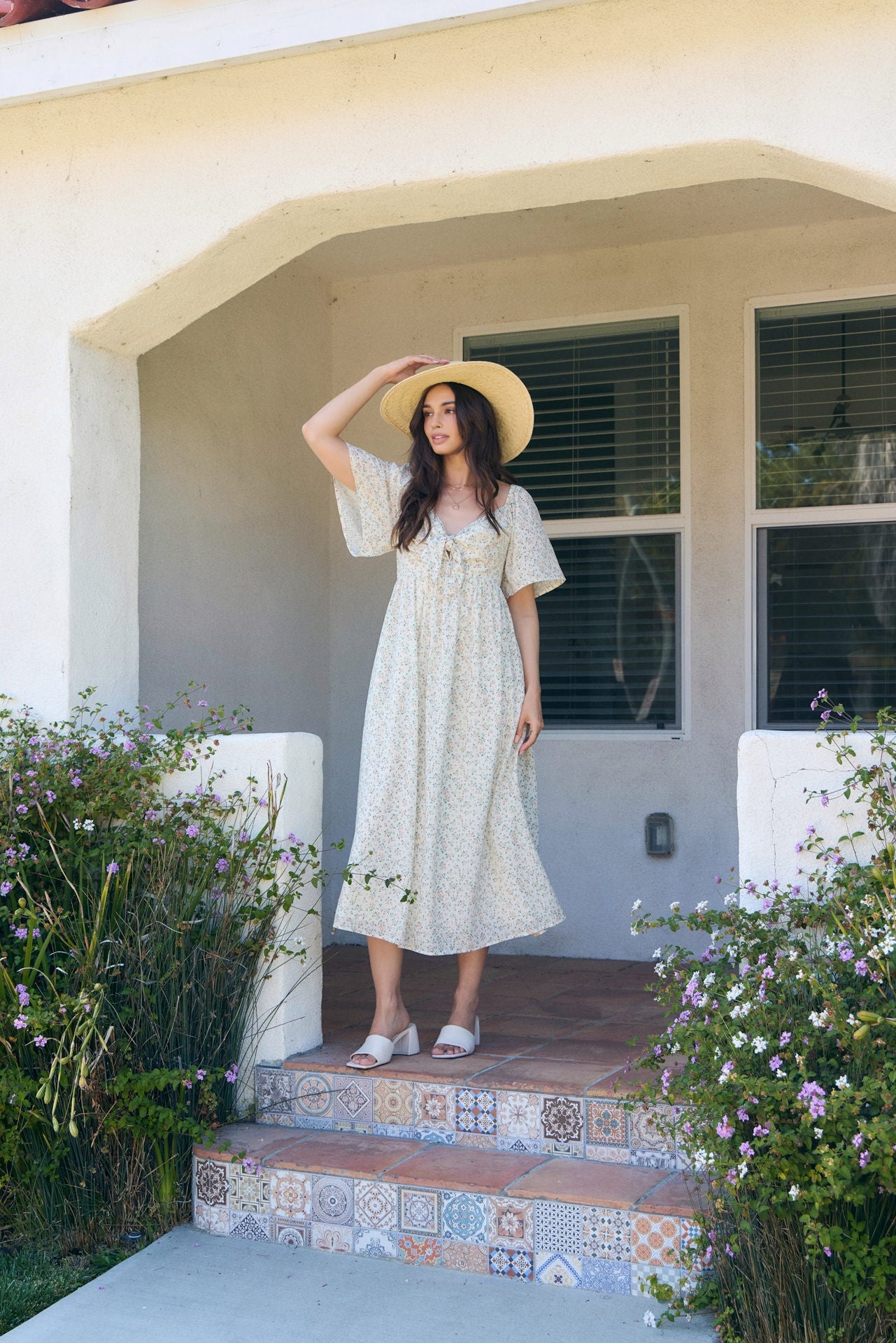 woman wearing a light floral maxi dress and a wide-brimmed hat, standing on a porch surrounded by greenery