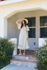 woman wearing a light floral maxi dress and a wide-brimmed hat, standing on a porch surrounded by greenery