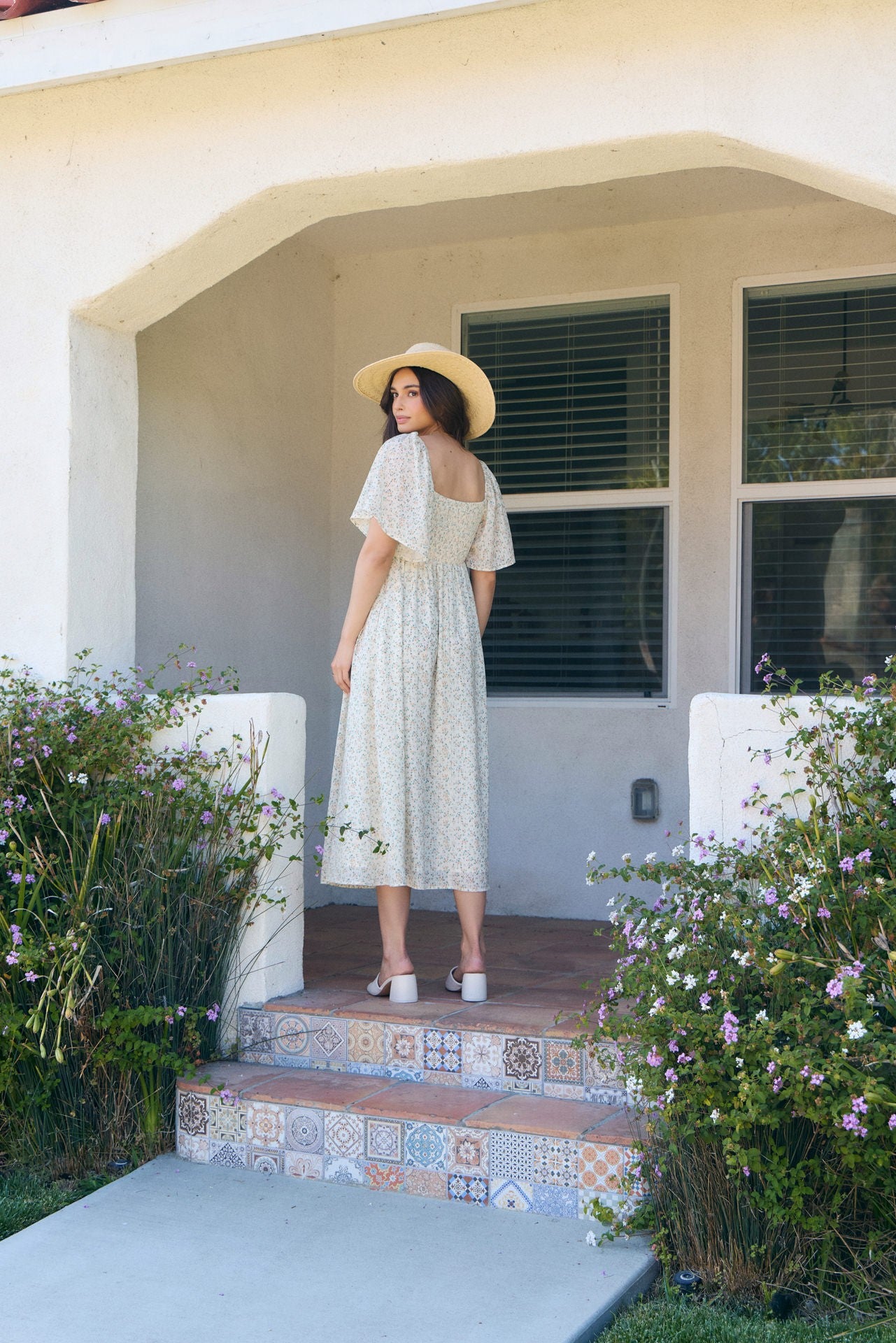 woman wearing a pale yellow floral midi dress with flutter sleeves and a wide-brimmed hat, standing on steps outside