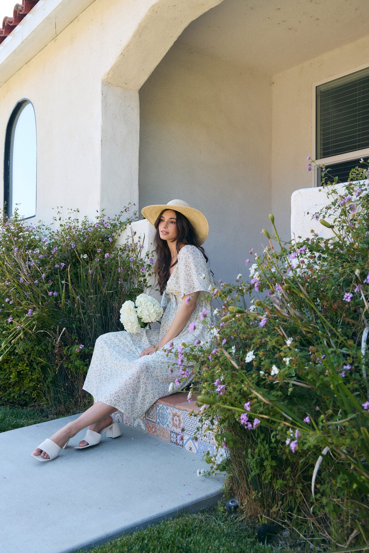 woman sitting on steps wearing a floral maxi dress and large straw hat, holding a bouquet of white flowers