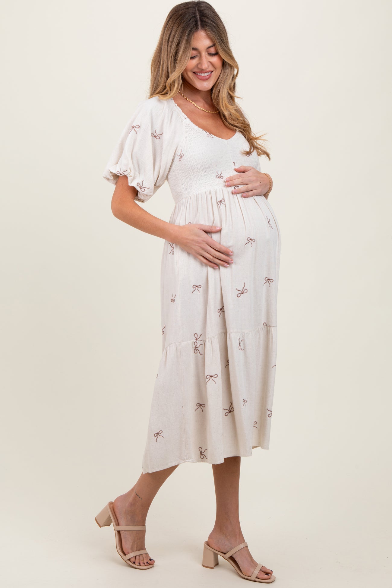 woman wearing a light beige maternity dress with short puff sleeves and bow print, cradling her baby bump