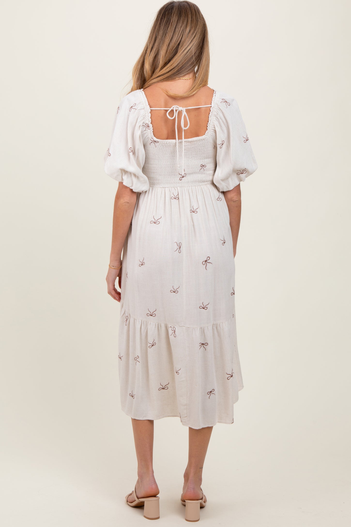 woman wearing a white maternity dress with puff sleeves and a tie back, featuring embroidered bows and a tiered hem