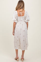 woman wearing a white maternity dress with puff sleeves and a tie back, featuring embroidered bows and a tiered hem