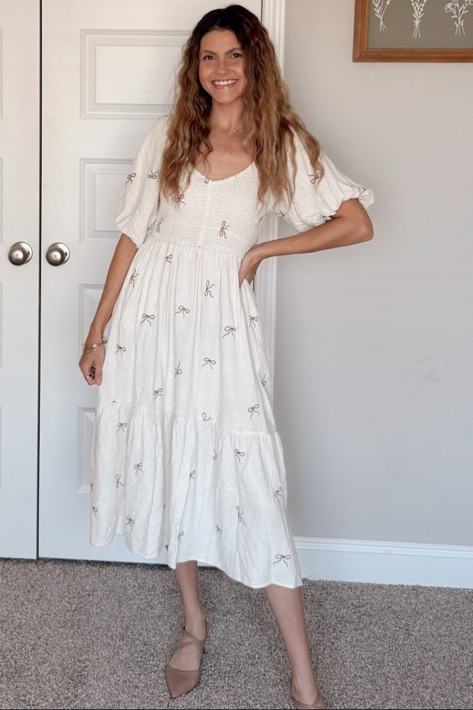 woman wearing a white midi dress with embroidered bow details and puff sleeves, standing in a stylish pose