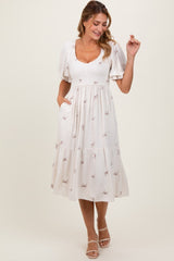 woman wearing a cream midi dress with puff sleeves and bow embroidery, smiling and posing with one hand raised