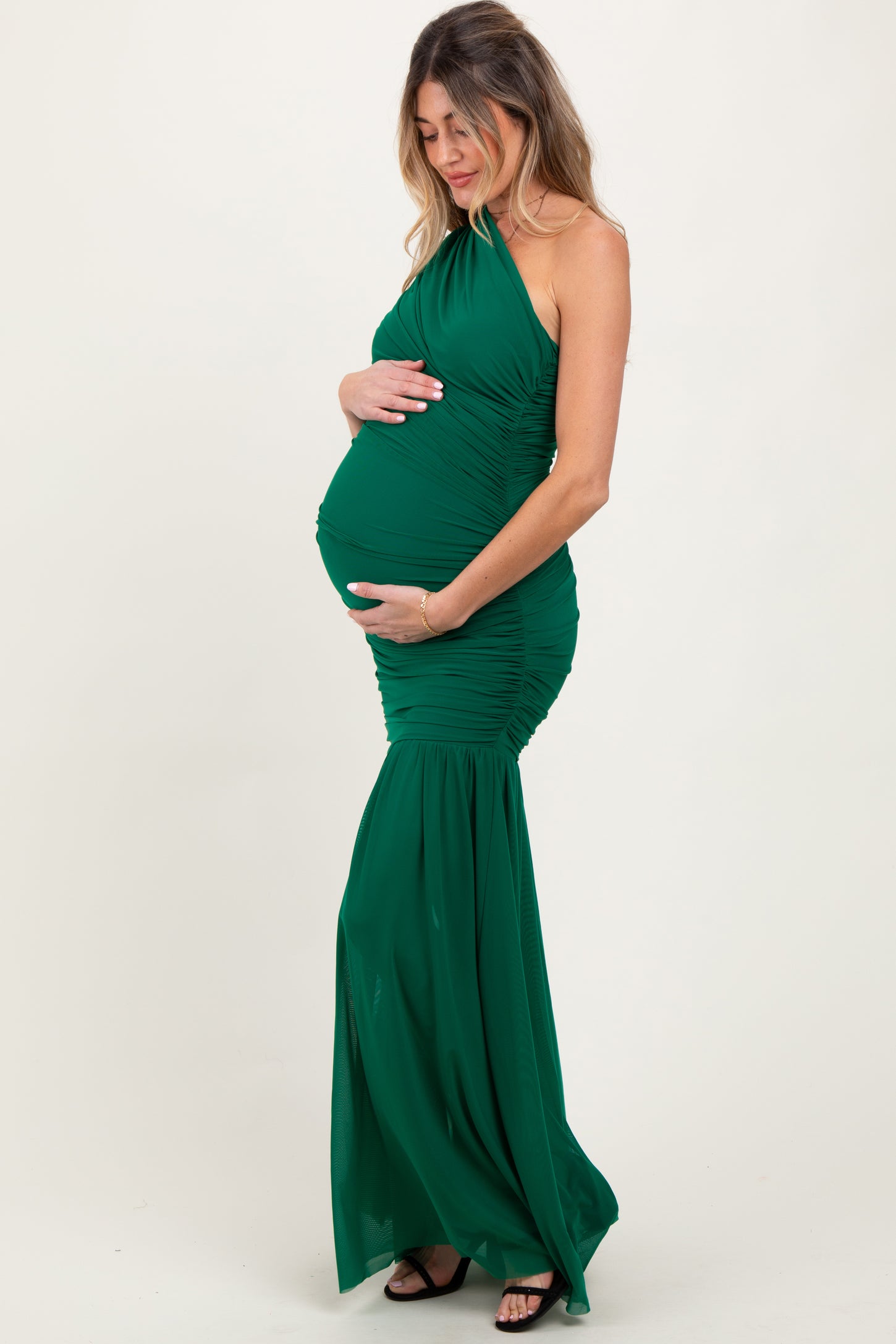 woman wearing a fitted green maternity gown with a halter neck and mermaid skirt, gently cradling her baby bump