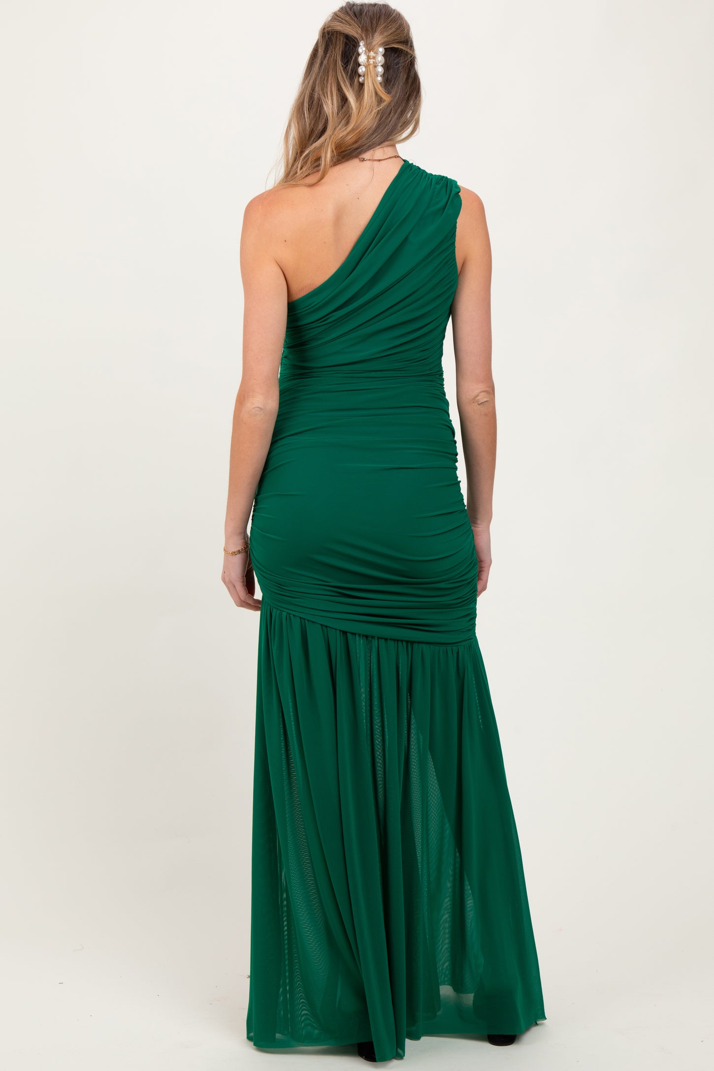 woman wearing a dark green one-shoulder maternity gown with ruching and a flowy skirt, showing her baby bump from behind