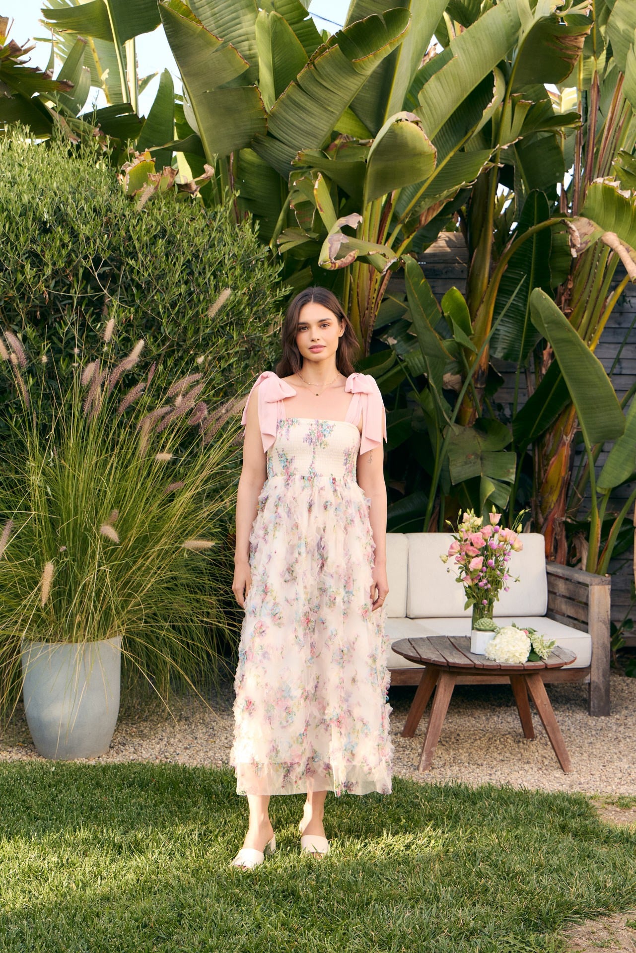 woman wearing a floral maxi dress with pink bow straps, standing in a garden with greenery and a floral arrangement nearby