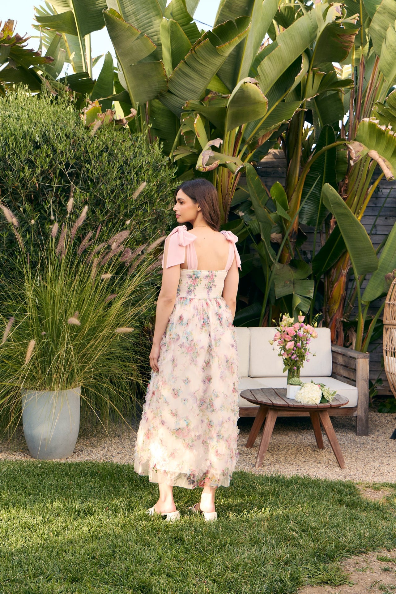 woman wearing a pink floral maxi dress with bow shoulder straps, standing outdoors surrounded by greenery