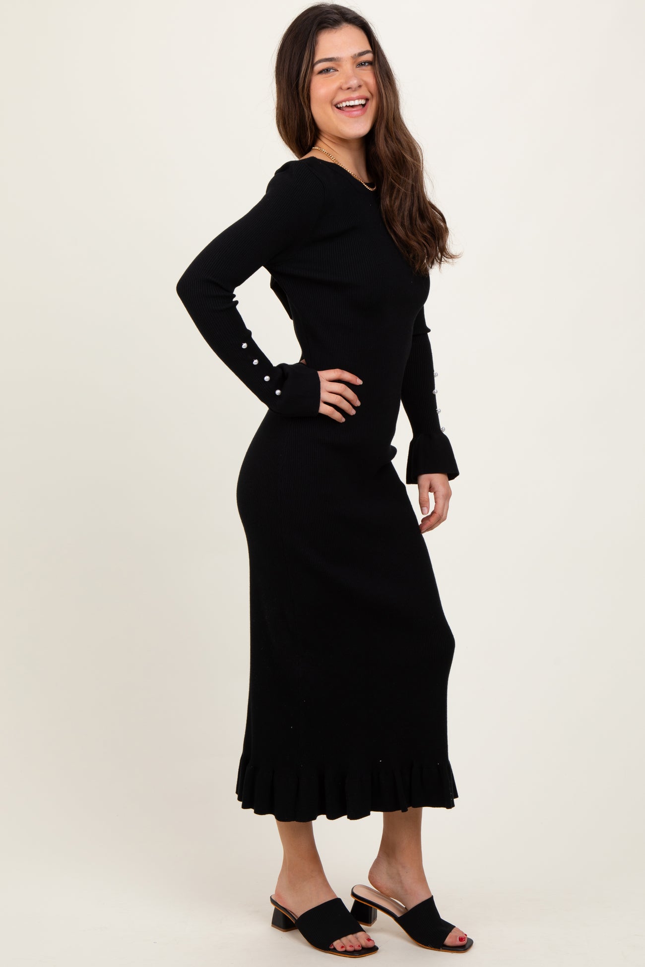 woman wearing a black ribbed long sleeve dress with ruffle hem and pearl button details, smiling and standing in profile