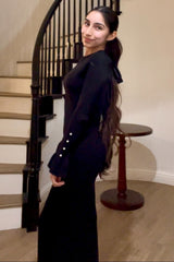 woman wearing a black long sleeve dress with a bow tie detail and button accents on the sleeves, standing by a staircase
