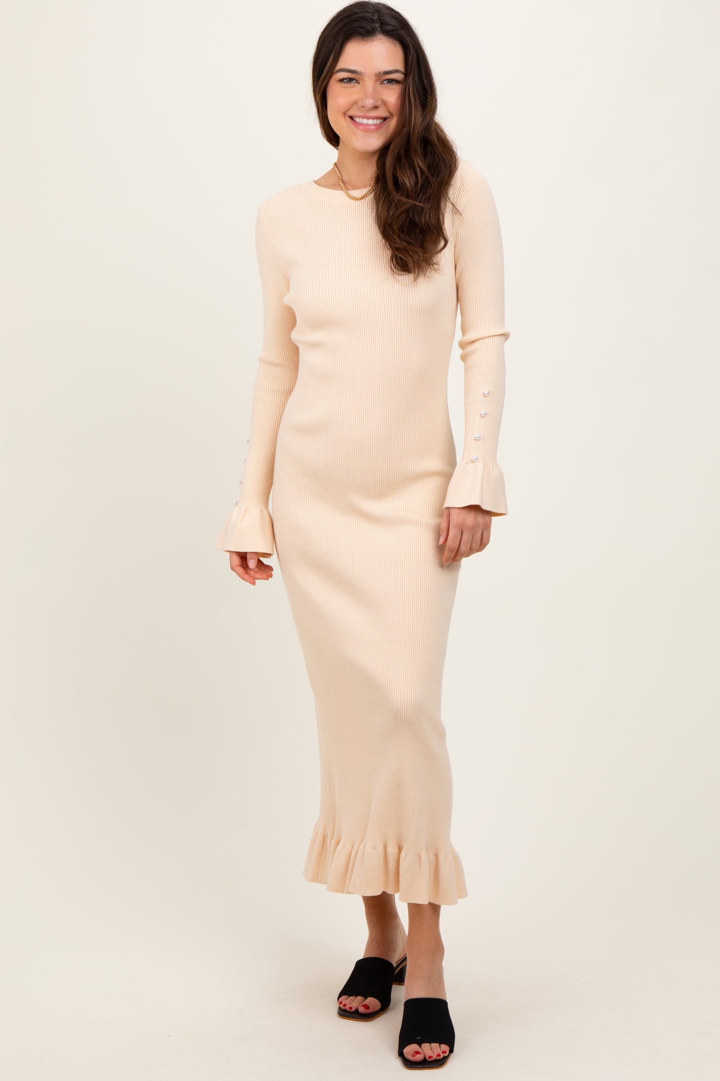 woman wearing a light beige ribbed long sleeve dress with bell sleeves and a ruffled hem, standing and smiling
