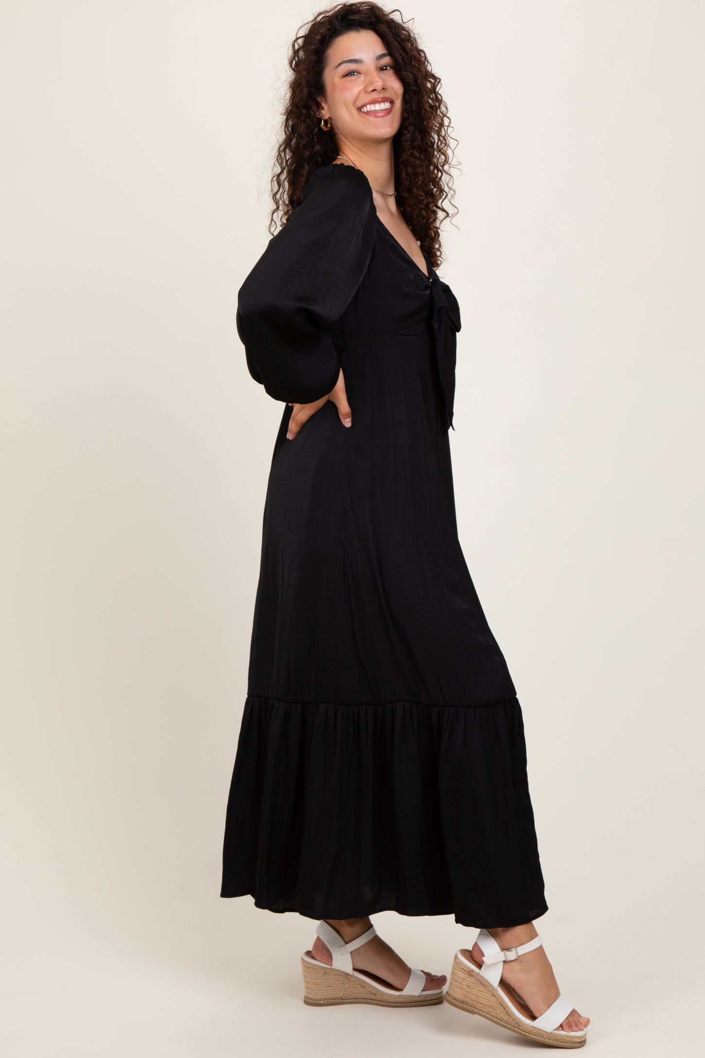woman wearing a black maxi dress with puff sleeves and a tied neckline, paired with white wedge sandals