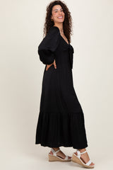 woman wearing a black maxi dress with puff sleeves and a tied neckline, paired with white wedge sandals