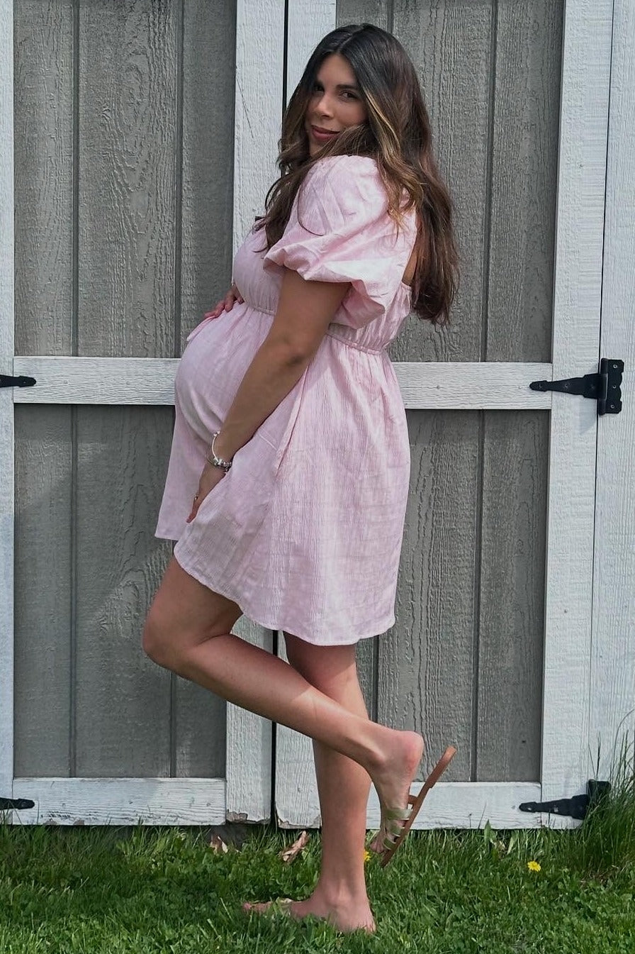 woman in a light pink short sleeve maternity dress showing her baby bump while standing next to a wooden fence