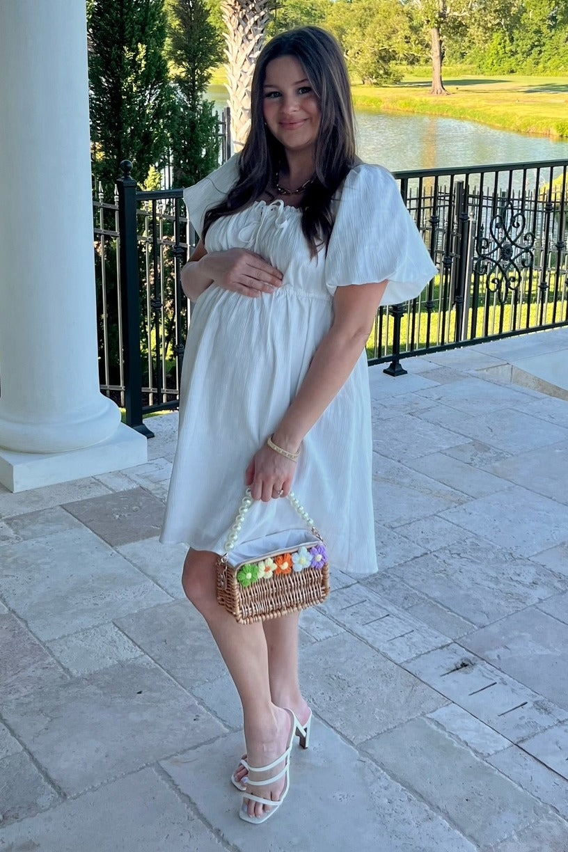 woman wearing a white maternity dress with puff sleeves, holding her baby bump and a basket of colorful eggs