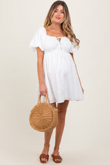 woman wearing a white maternity dress with puff sleeves and a tie detail, holding a straw handbag and showing her baby bump