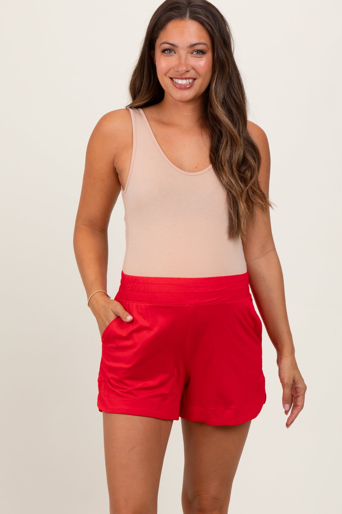 woman wearing a light tan tank top and red maternity shorts with pockets, smiling at the camera