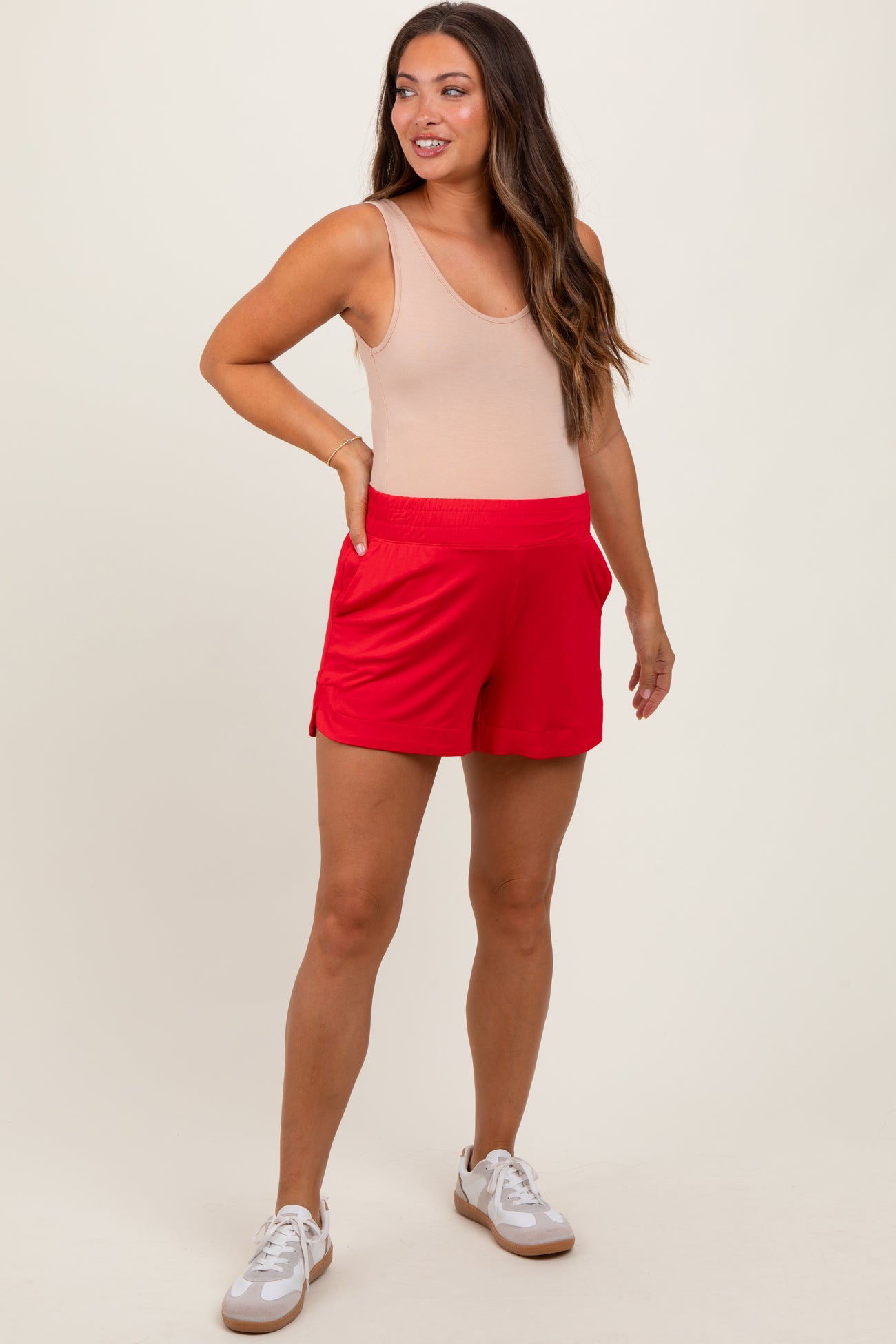 woman wearing a beige maternity tank top and red maternity shorts, smiling with one hand on her hip