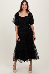 woman wearing a black puff sleeve midi dress with sheer overlay and ruffled hem, standing and smiling