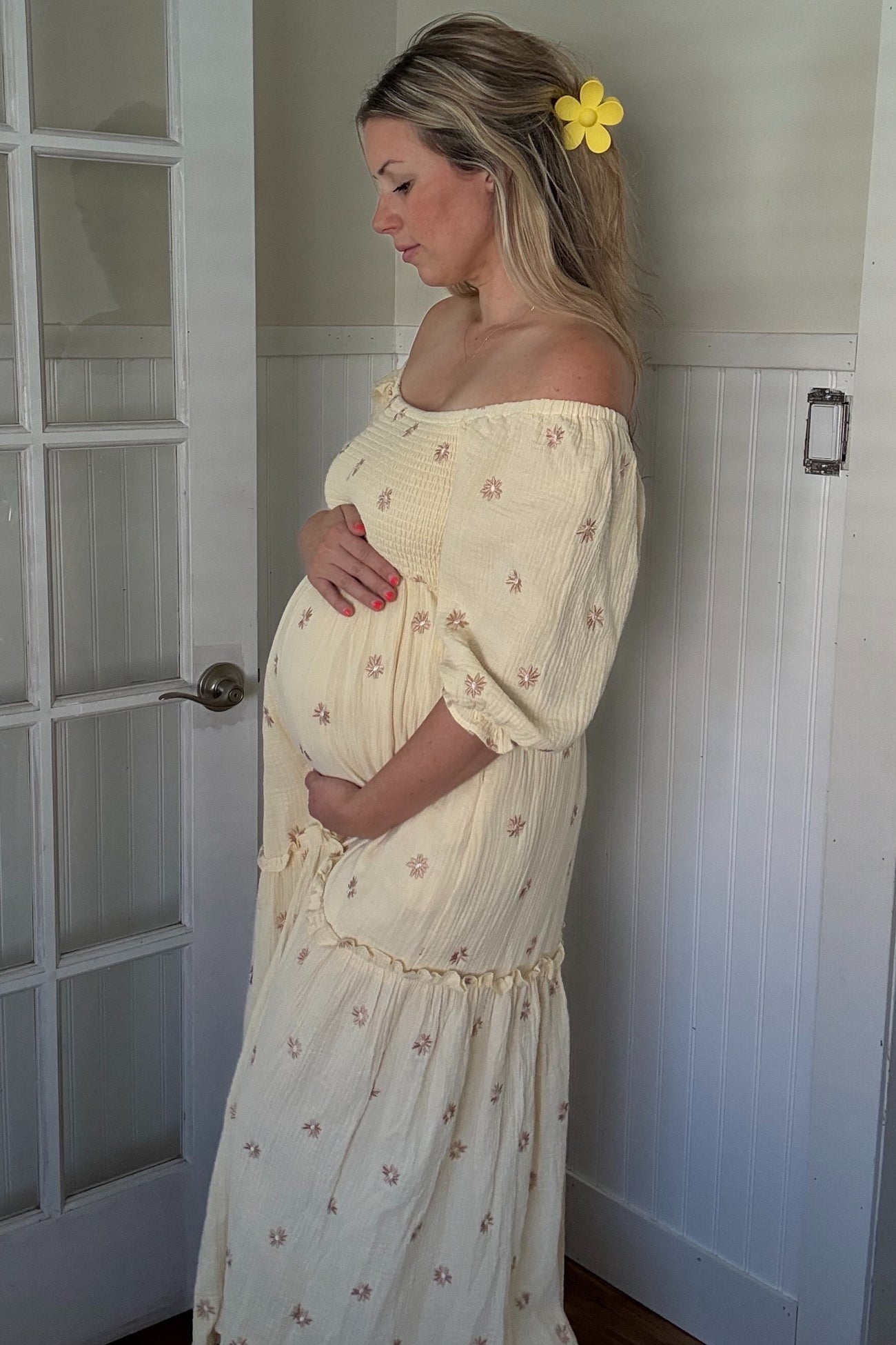 woman wearing a yellow off-shoulder maternity dress with floral details, gently cradling her baby bump