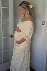 woman wearing a yellow off-shoulder maternity dress with floral details, gently cradling her baby bump