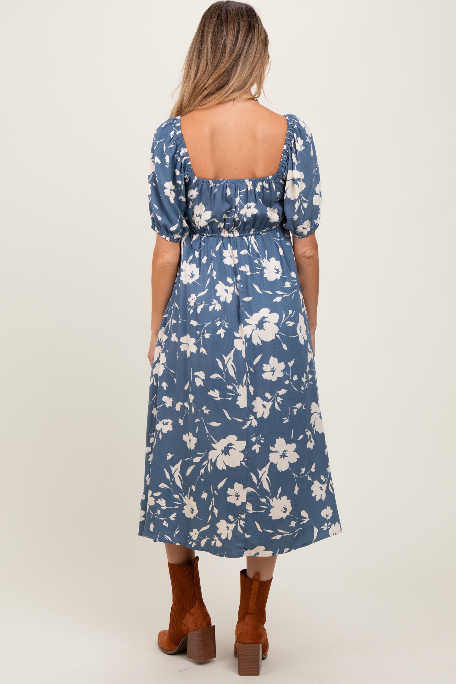 woman wearing a blue floral maternity dress with puff sleeves and a back cutout, styled with brown ankle boots