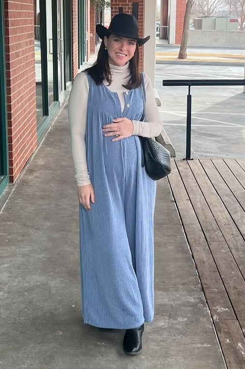 woman wearing a light blue maternity jumpsuit over a cream turtleneck and holding her baby bump