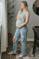 woman wearing a light green maternity tank top and distressed light blue jeans, cradling her baby bump