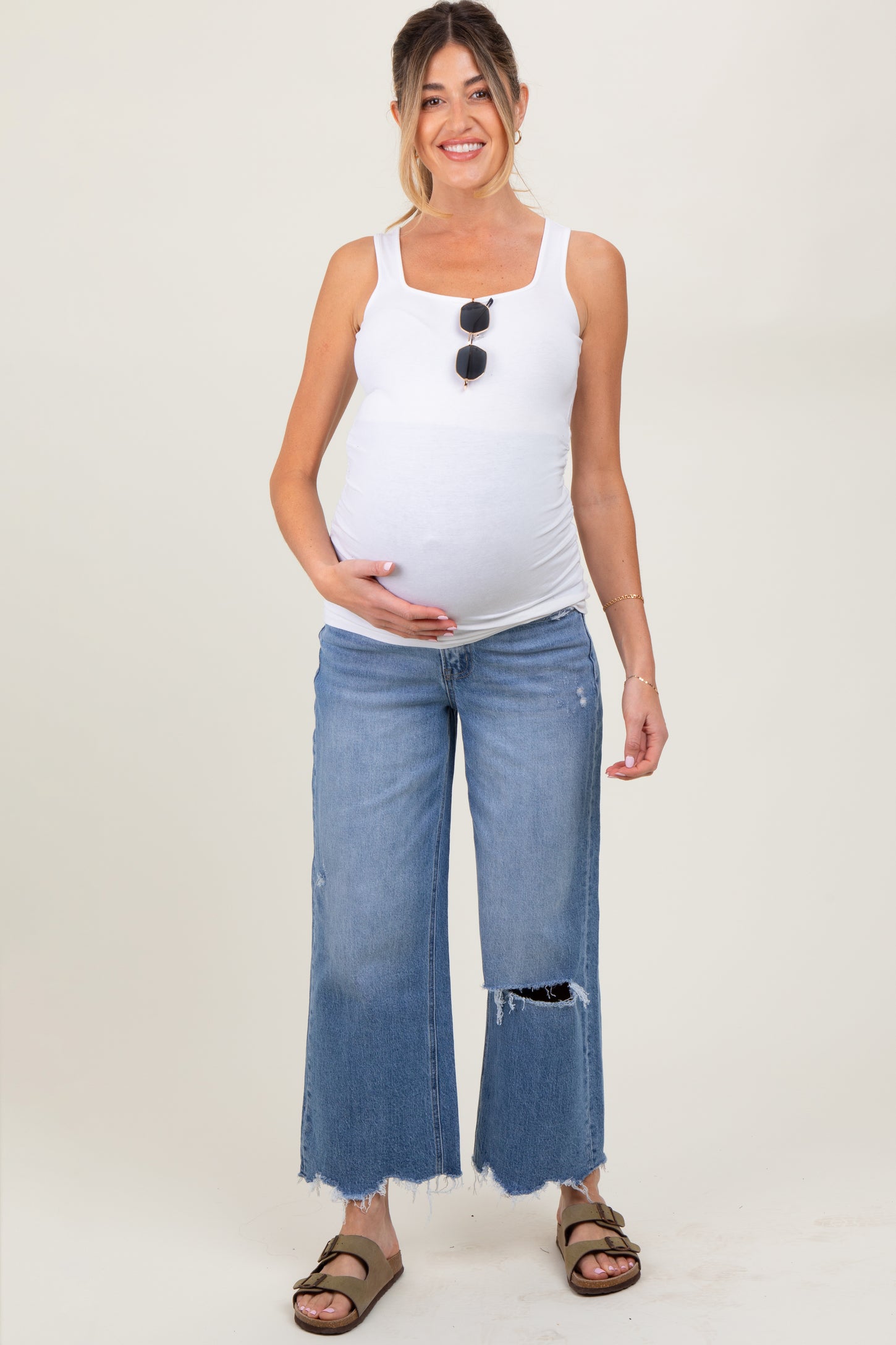 woman wearing a white maternity tank top and distressed wide-leg jeans, smiling and holding her baby bump