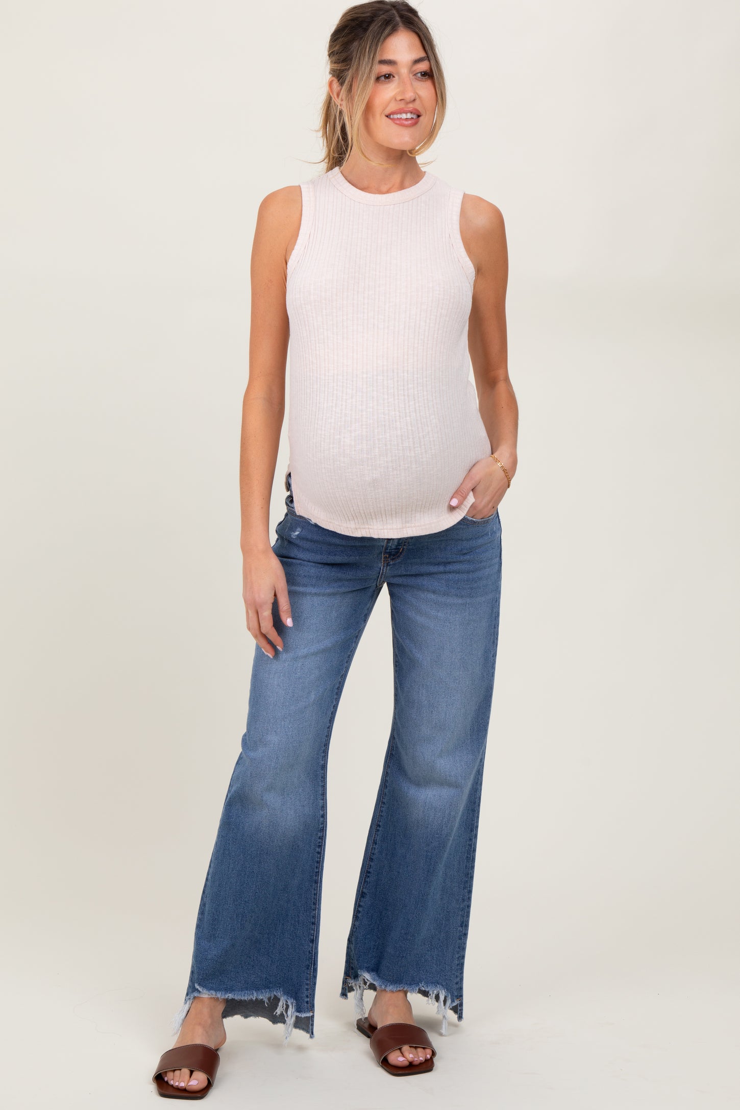woman wearing a light pink sleeveless maternity top and blue flared jeans, showing her baby bump