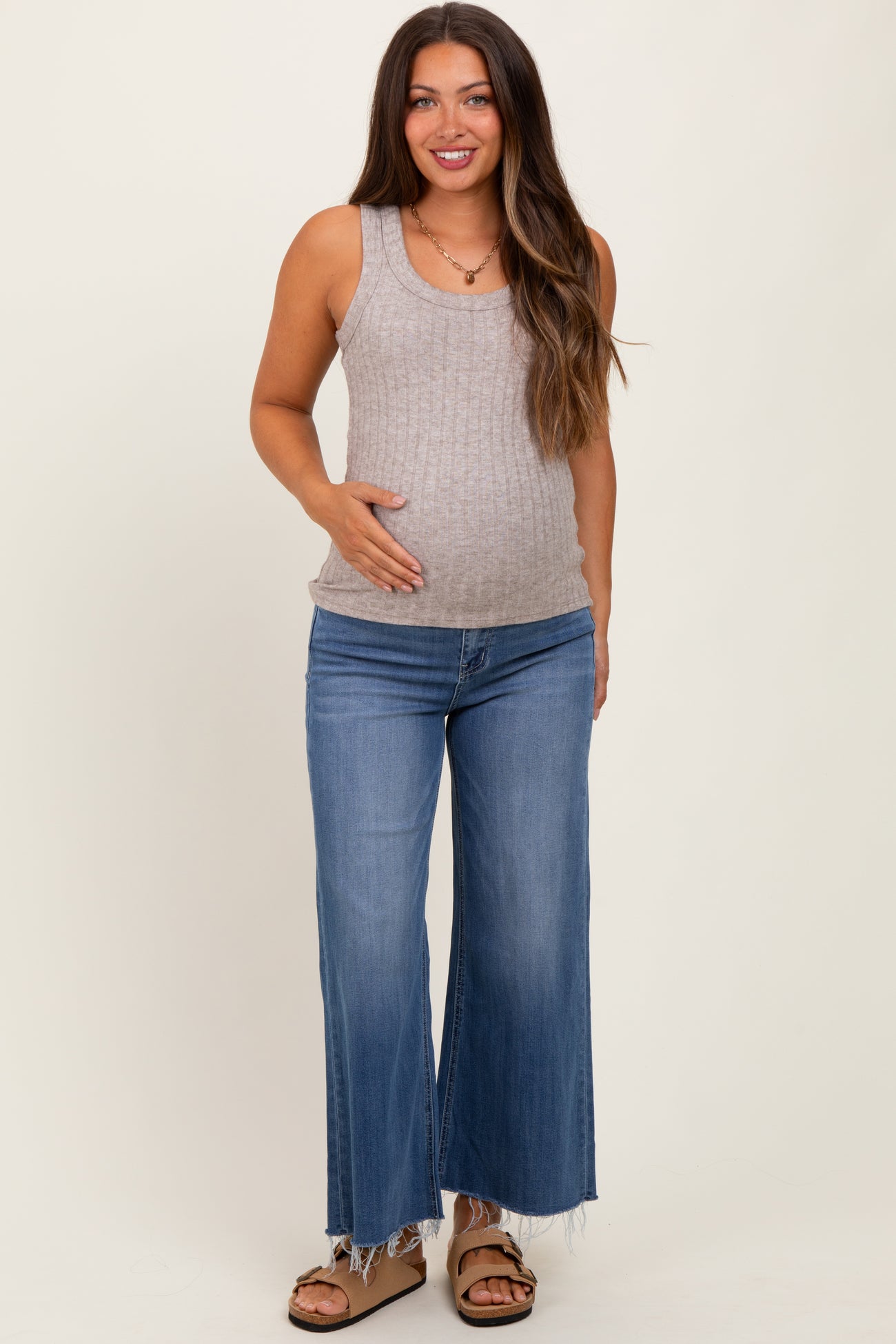 woman wearing a light gray ribbed maternity tank top and distressed wide-leg jeans, gently holding her baby bump