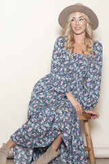 woman wearing a floral long sleeve maxi dress and a wide-brimmed hat, sitting on a stool