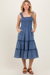 woman wearing a denim blue tiered midi dress with smocked bodice and ruffled shoulder straps, posing with hands in pockets