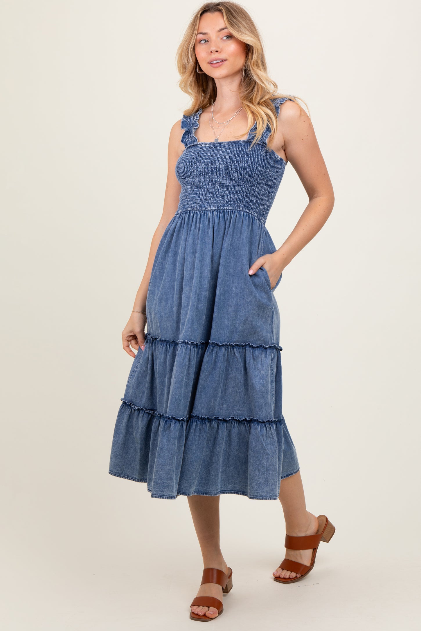 woman wearing a denim smocked midi dress with ruffled tiers and pockets, standing on a neutral background