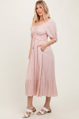 woman wearing a blush pink midi dress with puff sleeves and a ruched bodice, featuring a tiered skirt and pockets