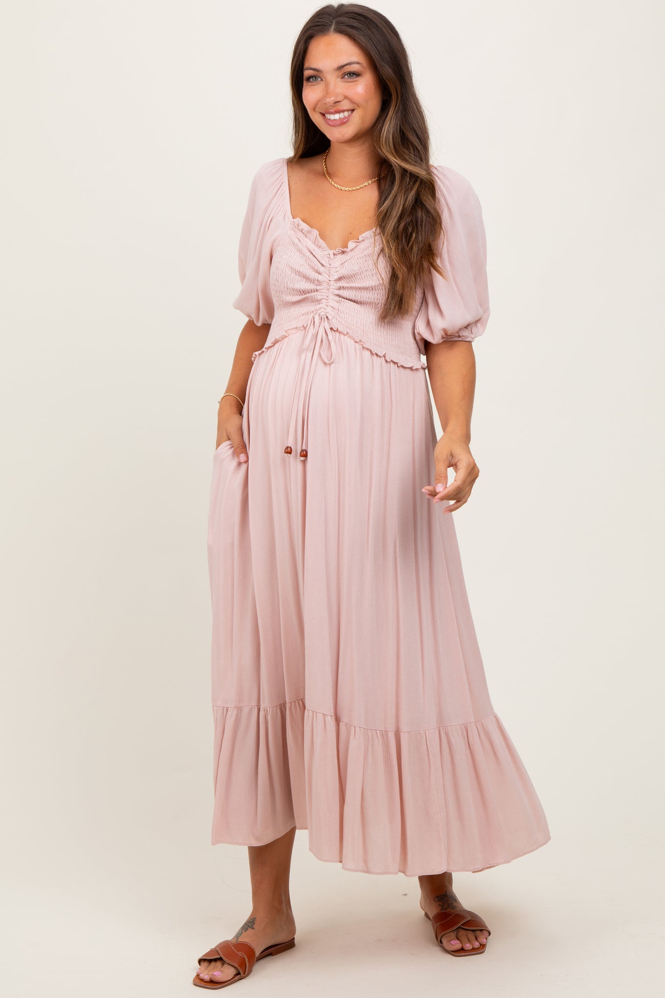 woman wearing a blush pink maternity maxi dress with puff sleeves and a smocked bodice, smiling and posing with hands in pockets