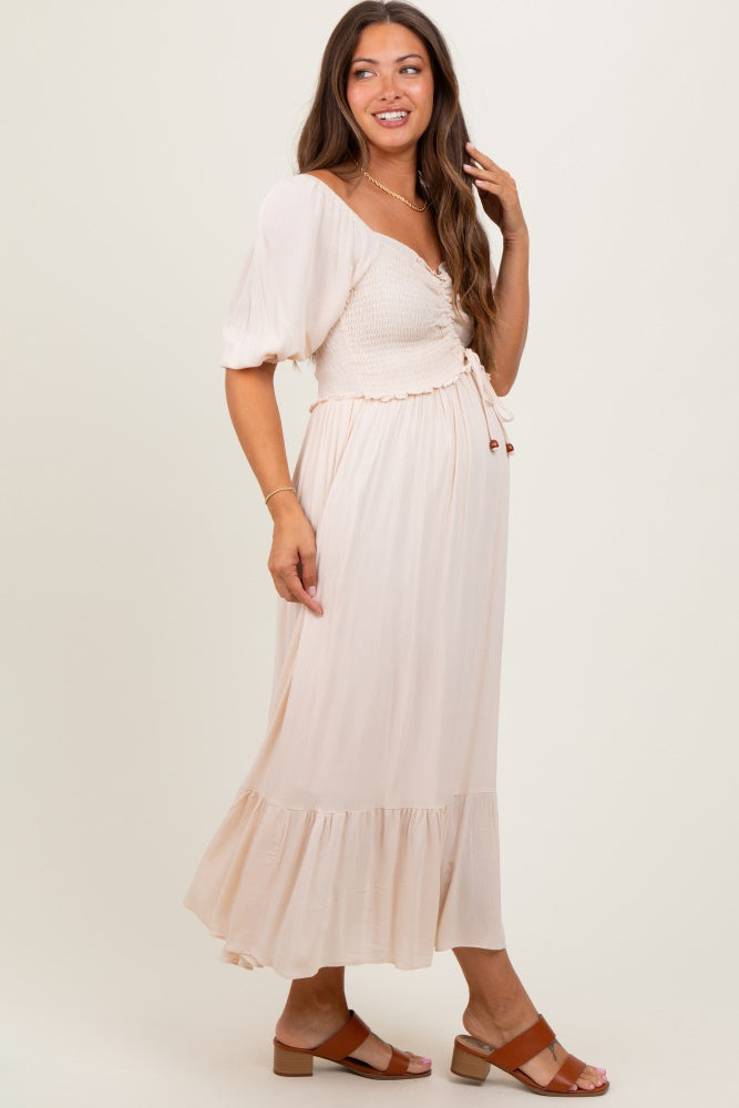 woman wearing a light beige maternity dress with a smocked bodice and flared hem, looking at the camera with a smile