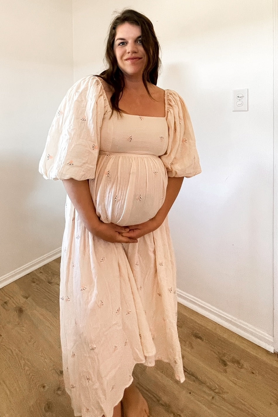 woman wearing a light pink maternity dress with puffy sleeves and floral embroidery, holding her baby bump